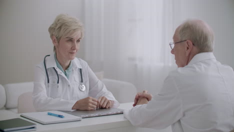 elderly-male-therapist-is-consulting-with-woman-head-physician-of-hospital-in-office-discussing-and-deciding